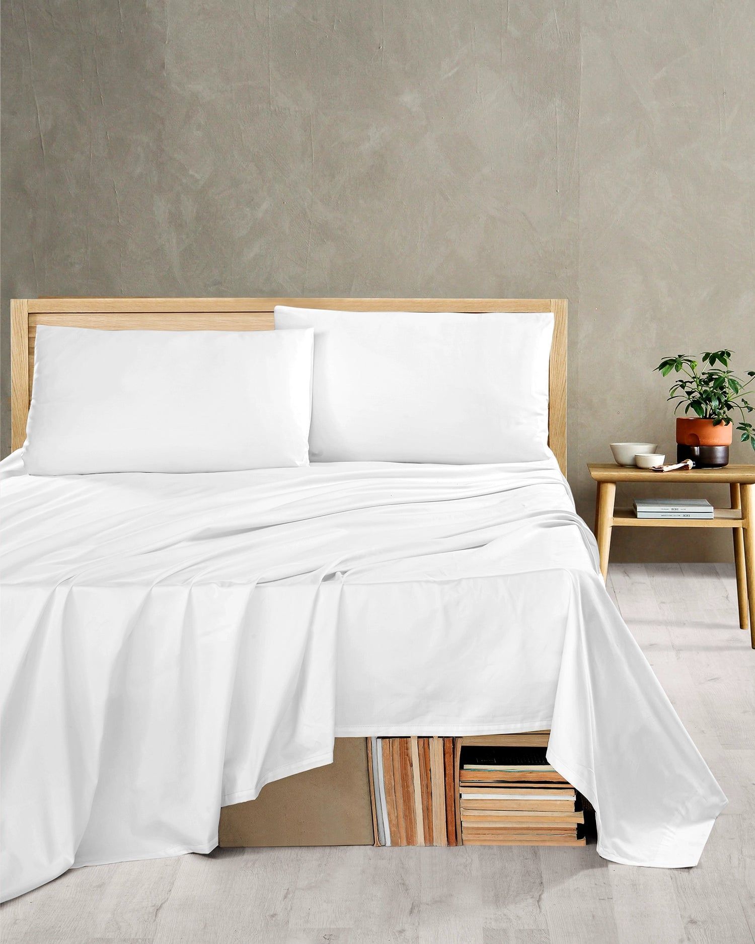 White bed with rumpled bedding in a minimalist bedroom, beside a wooden nightstand with a plant and books.