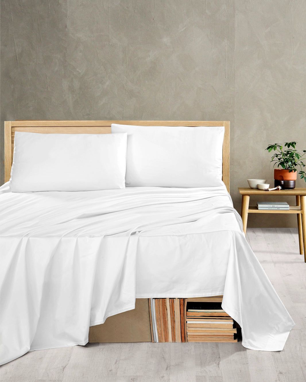 White bed with pillows and draped sheets in a minimalist bedroom beside a wooden nightstand and plant