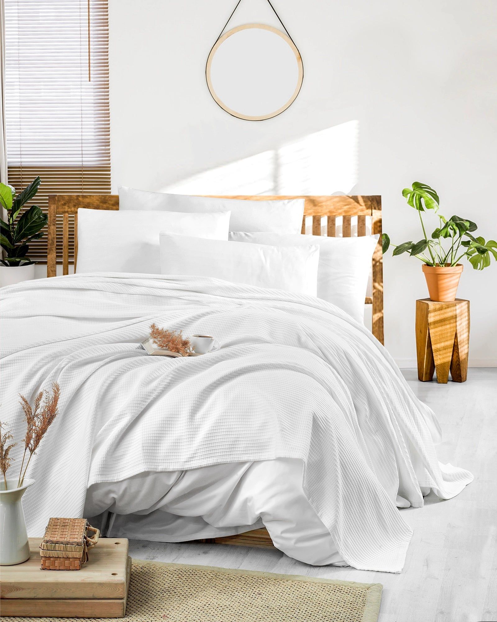 Bright white bedroom with unmade bed, wooden accents, plants, and natural light through a window.
