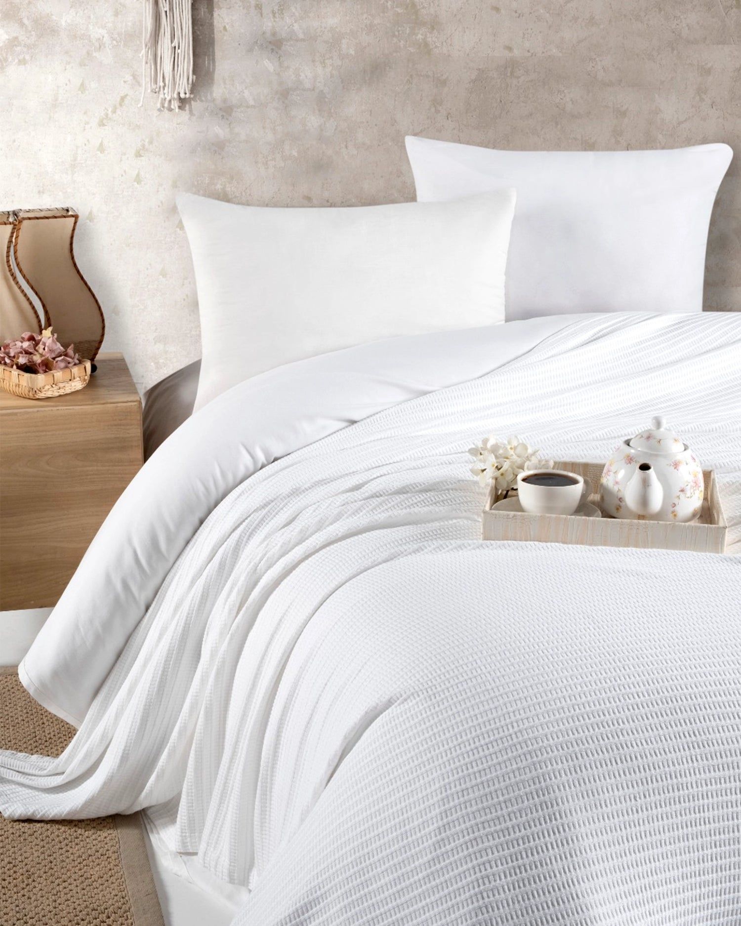 White bed with textured bedding and pillows, with a tray and decor on a warm-toned bedside table.