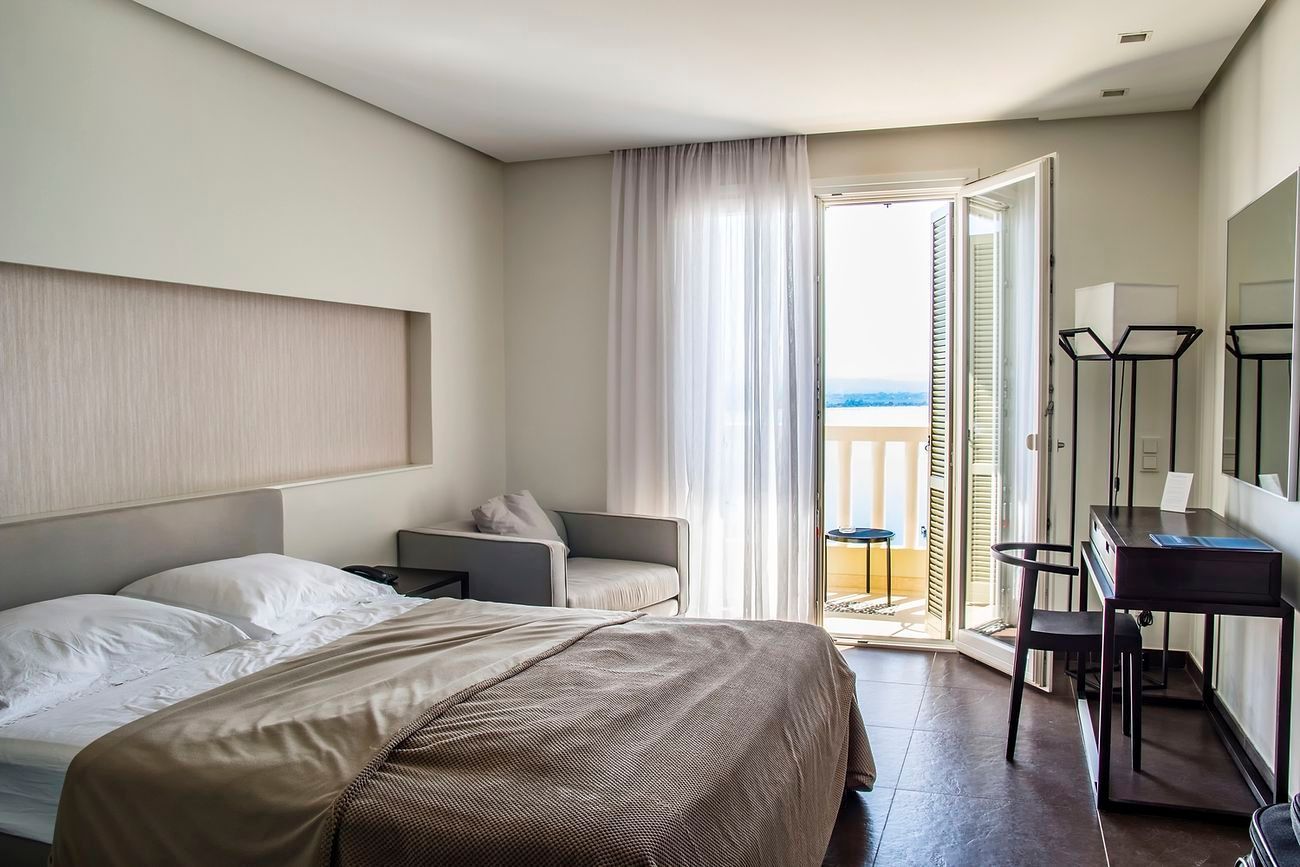 Bright bedroom with bed, sofa, desk, and open balcony doors letting in daylight.