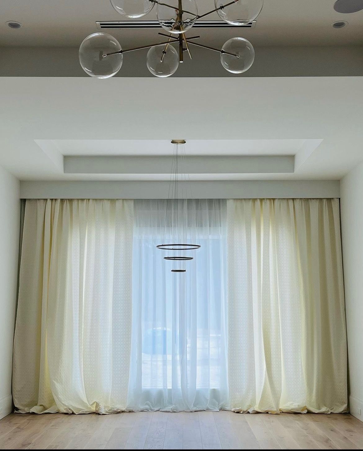 Empty room with sheer white curtains, pale walls, and a modern chandelier near the ceiling.