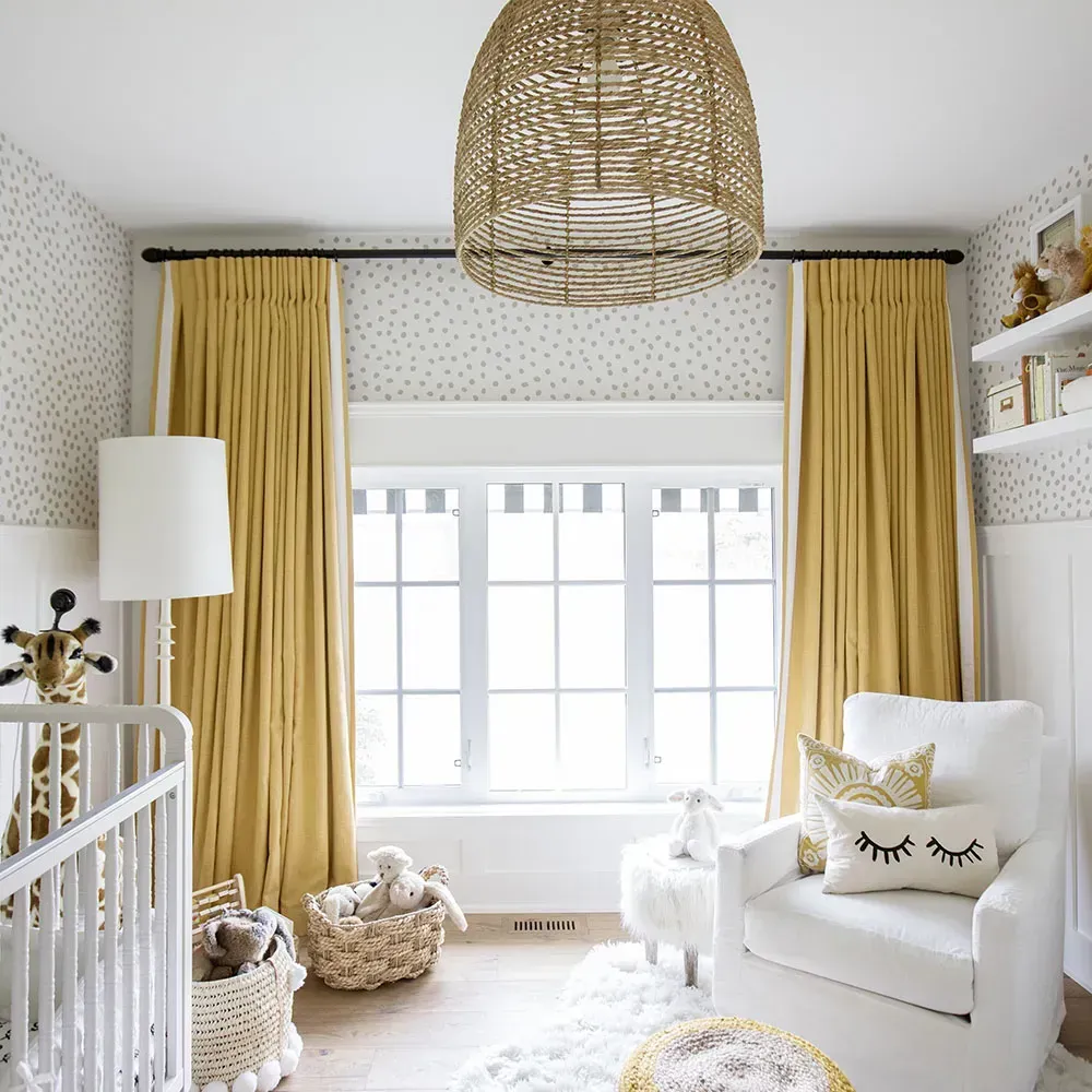 Bright nursery with yellow curtains, white armchair, wicker pendant light, and a window wall.