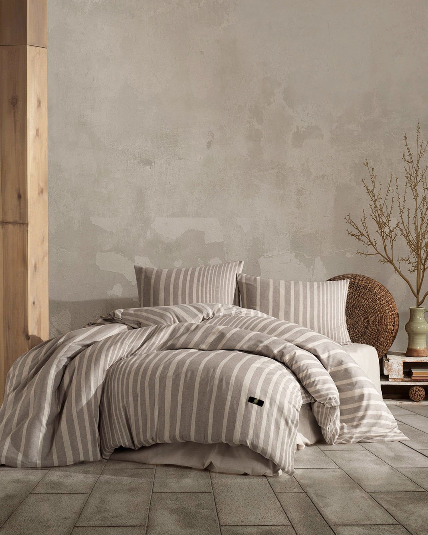 Striped bedding on an unmade bed in a minimalist beige bedroom with a woven vase and dried branches