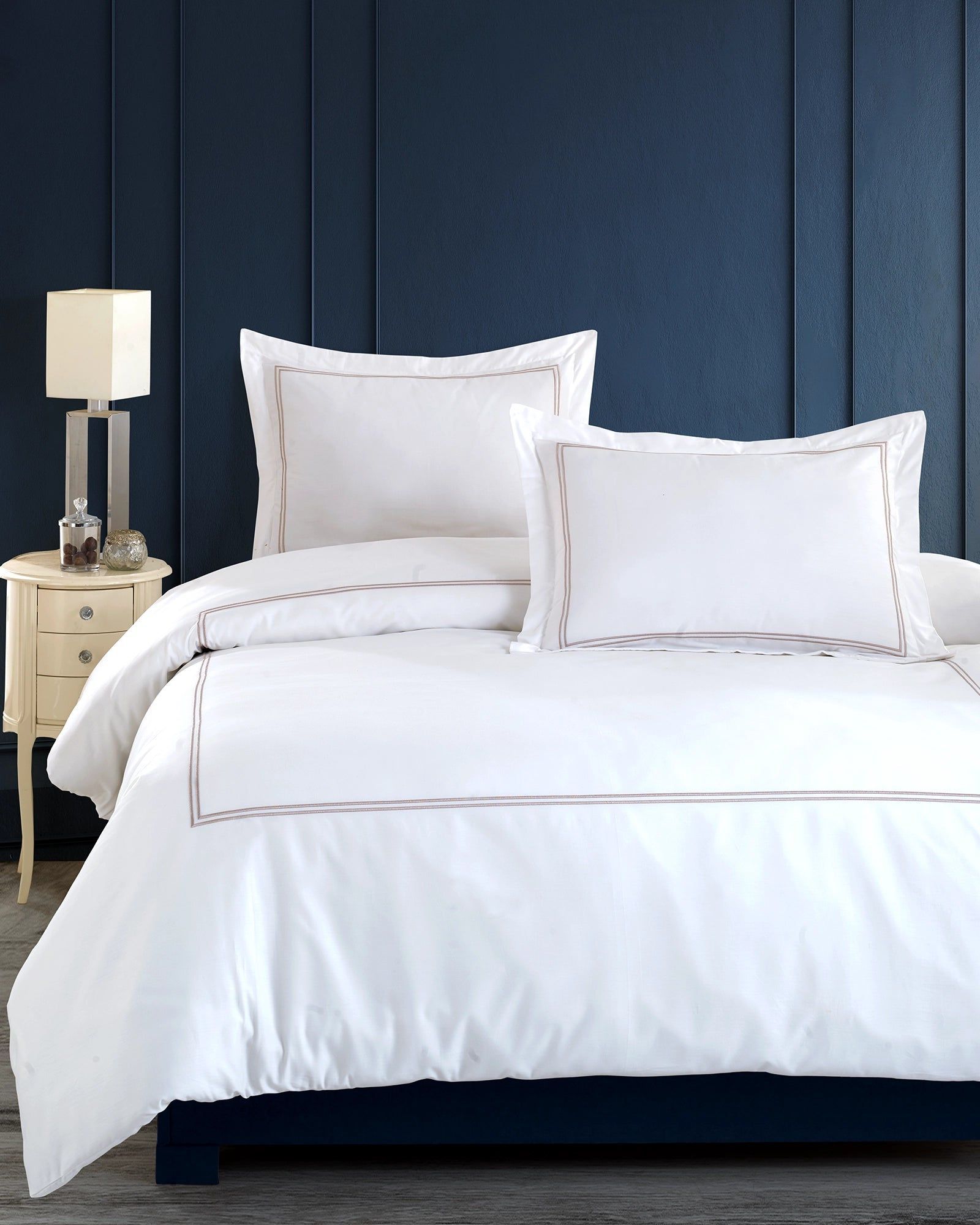 White bed with pillows and duvet in a blue bedroom beside a small nightstand lamp.