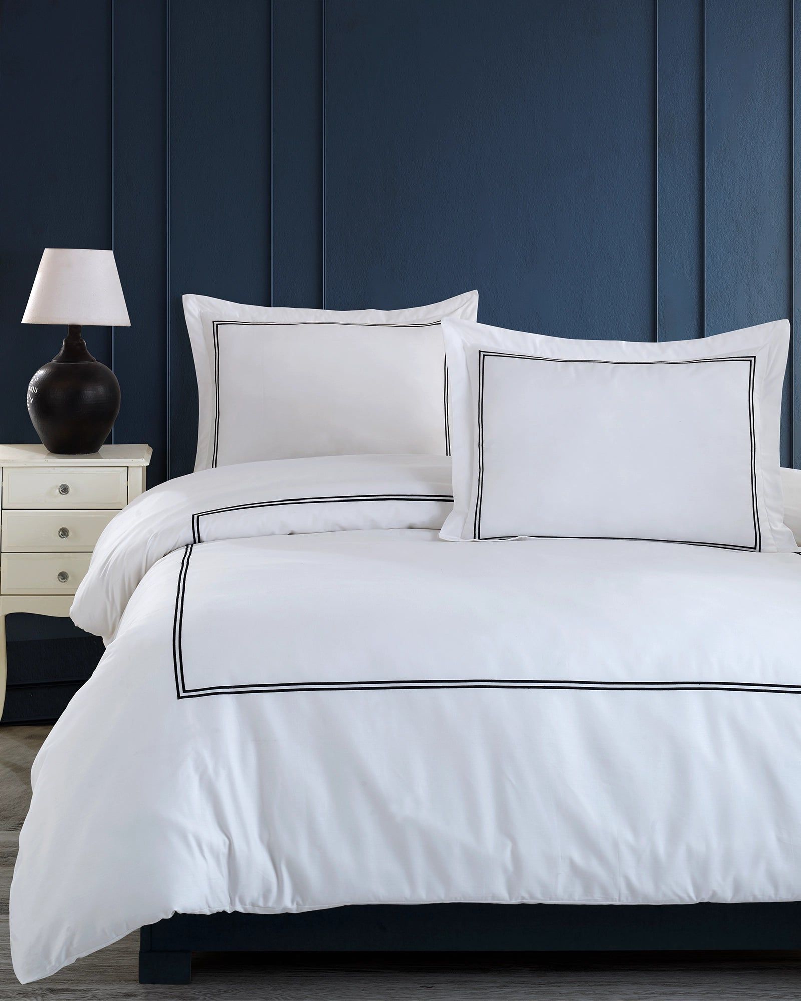 White bed with navy throw pillows and duvet in a dark blue bedroom, beside a small lamp on a nightstand