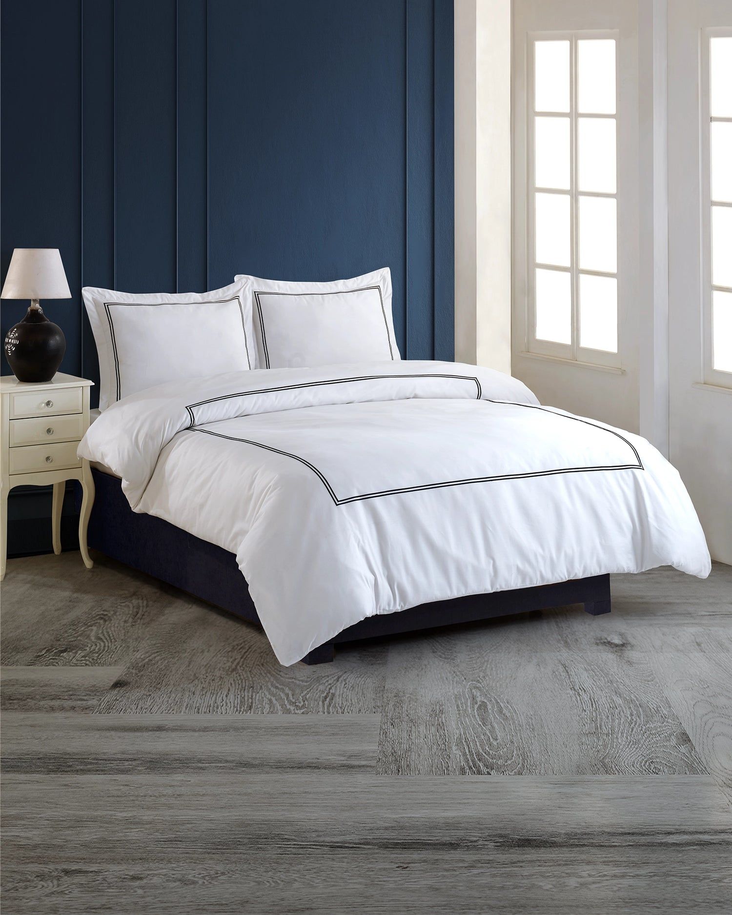 White bed with black trim in a blue bedroom with a nightstand and windows