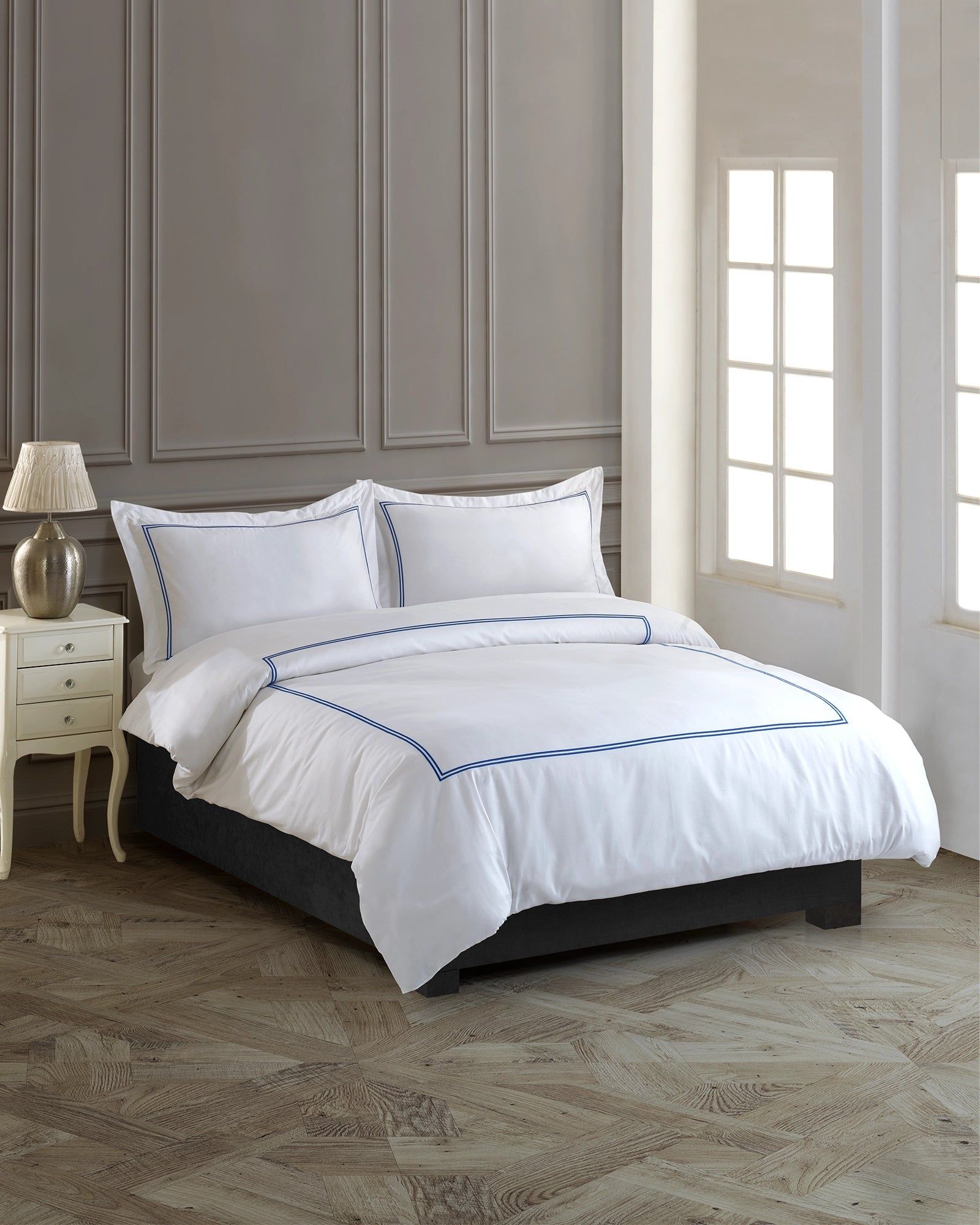 White bed with gray bedding in a elegant bedroom beside a window and nightstand lamp
