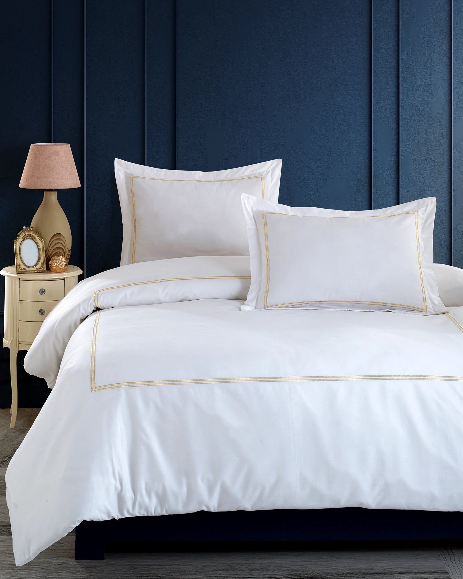 White bed with pillows and gold trim in a navy blue bedroom with a bedside lamp and clock