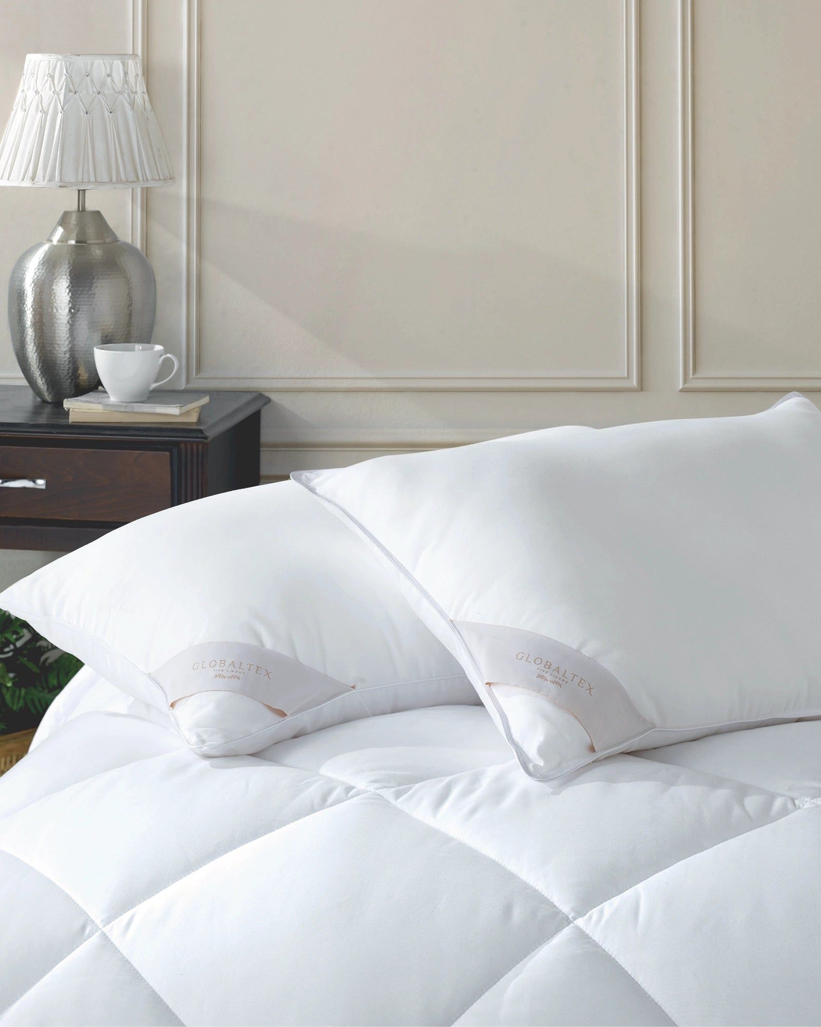 White bedding with pillows in a softly lit bedroom beside a lamp and nightstand