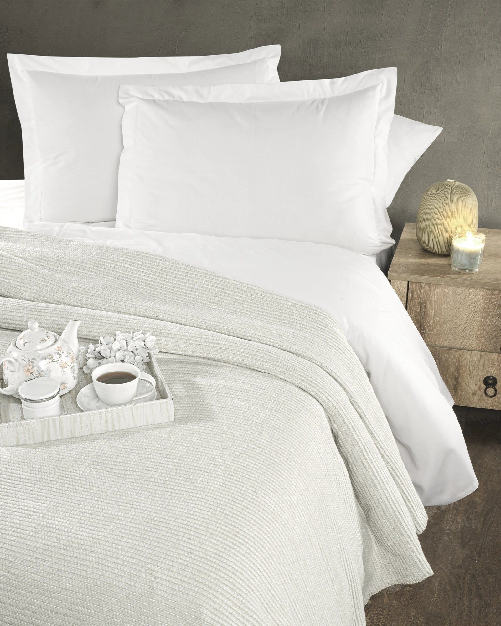 White bedding with pillows and a knit blanket on a bed beside a nightstand with a lamp.