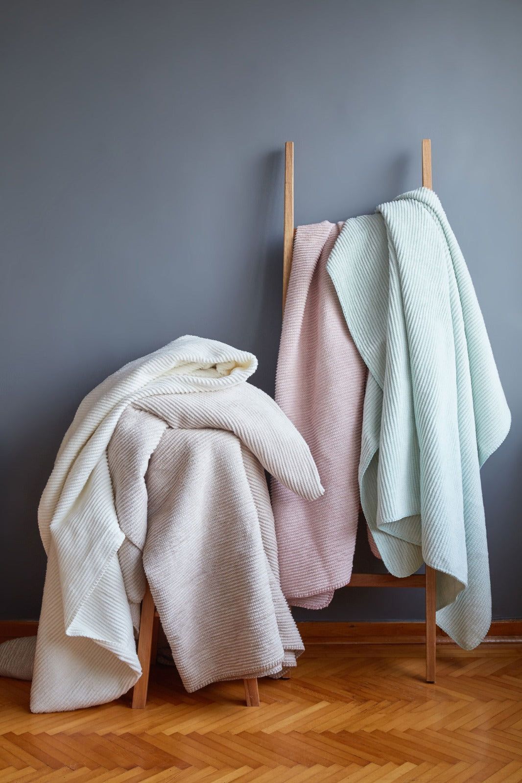 Three knit blankets draped over wooden stands against a gray wall and parquet floor