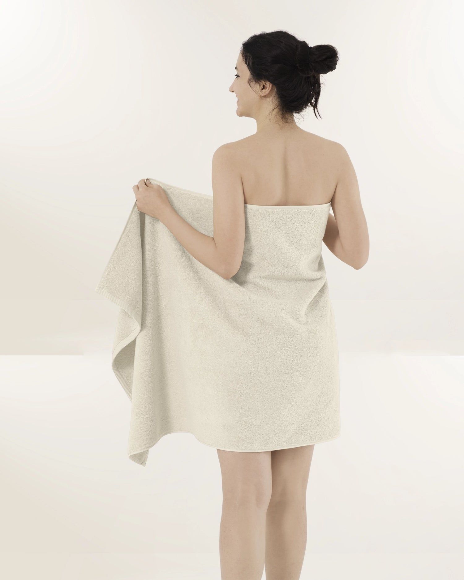 Woman wrapped in a light beige towel, seen from behind against a plain white background.