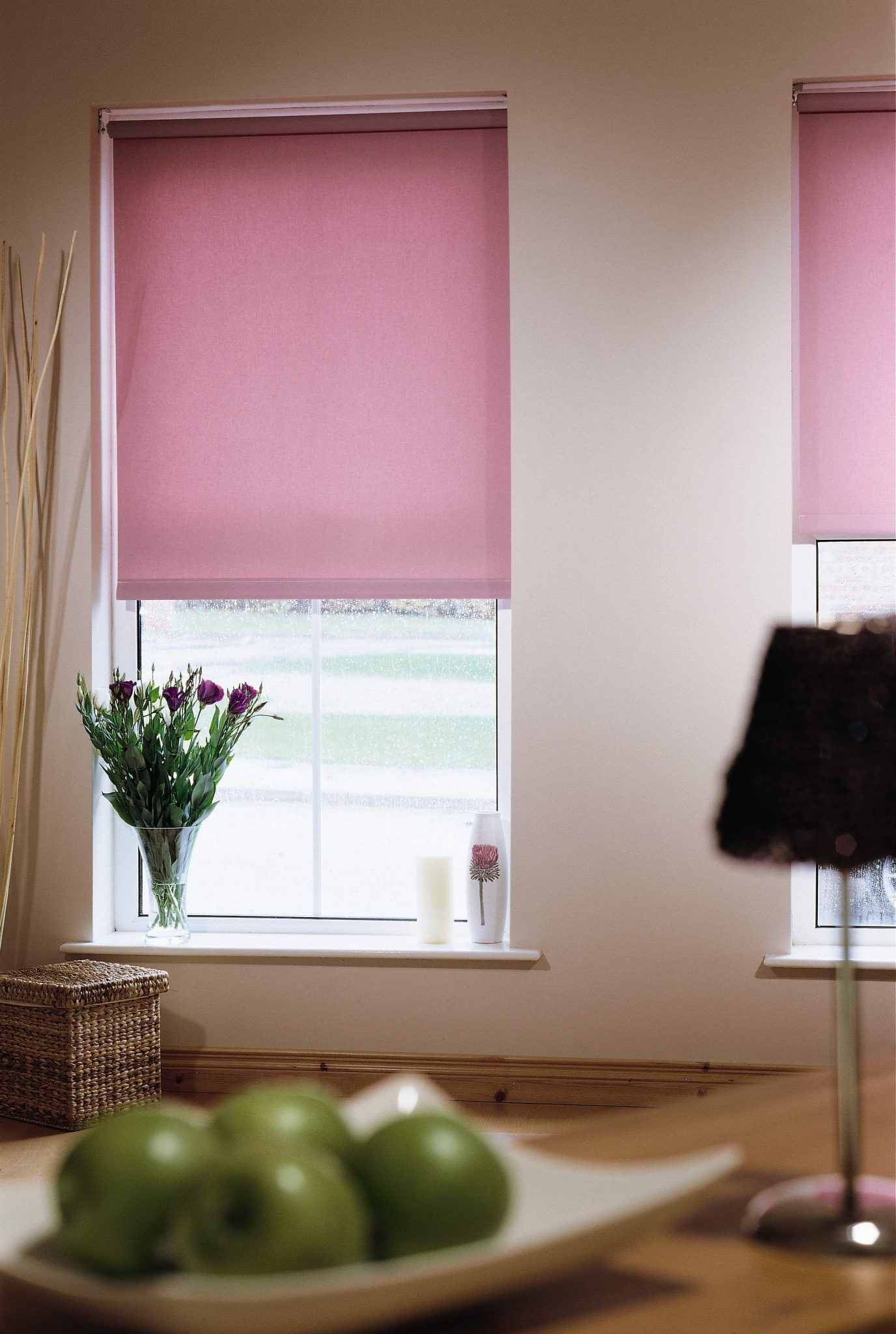 Pink blinds over a window with flowers on the sill and green apples on a table in the foreground