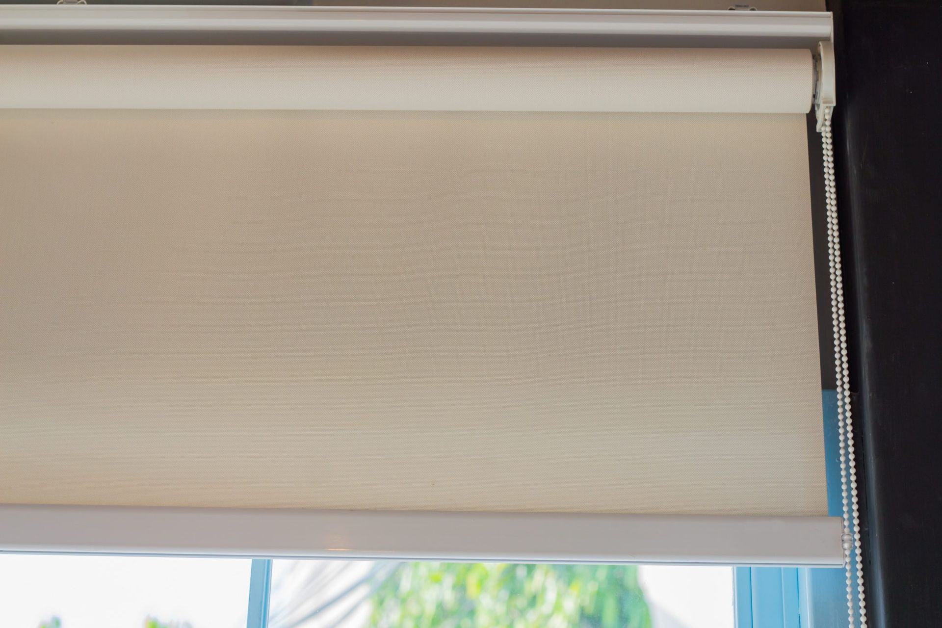 Beige roller blind partially closed over a window with greenery visible below