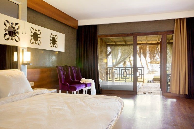 Modern hotel room with white bed, purple chair, and balcony doors overlooking trees