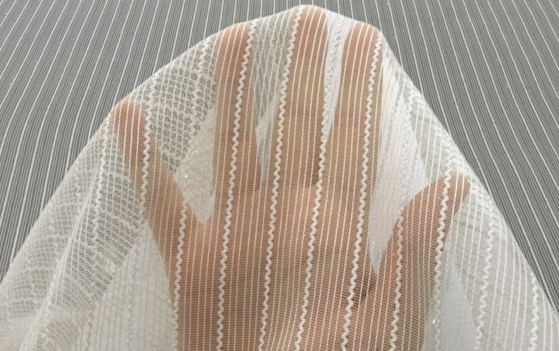 Hand behind sheer white lace curtain, with beige striped pattern over gray woven background