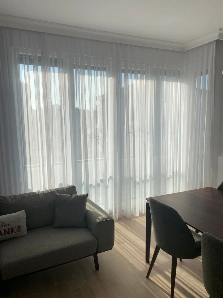 Bright living room with sheer white curtains, gray sofa, and dark dining table by a sunlit window.