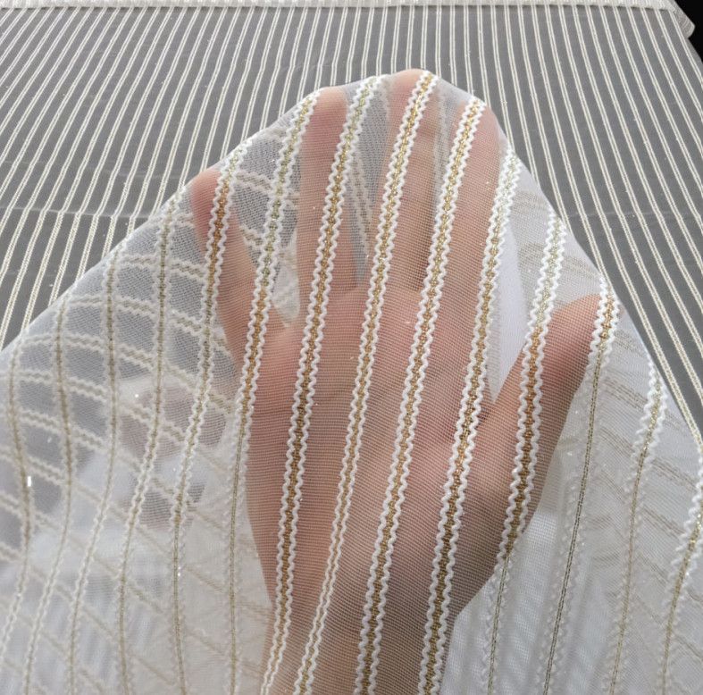 A hand behind white mesh fabric with gold and pink stripes against a gray patterned background