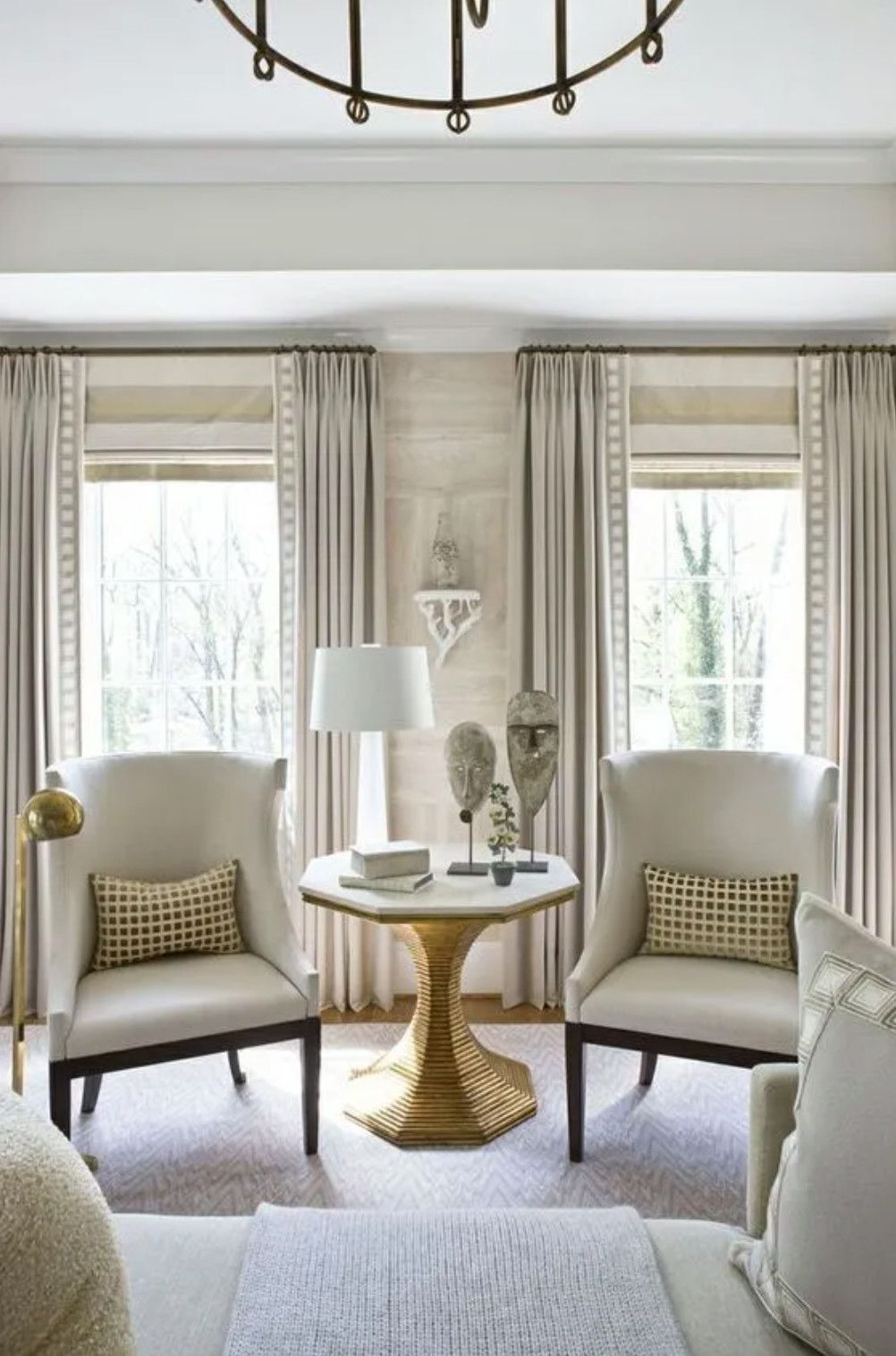 Elegant living room with two beige armchairs, gold side table, lamp, and tall windows with curtains