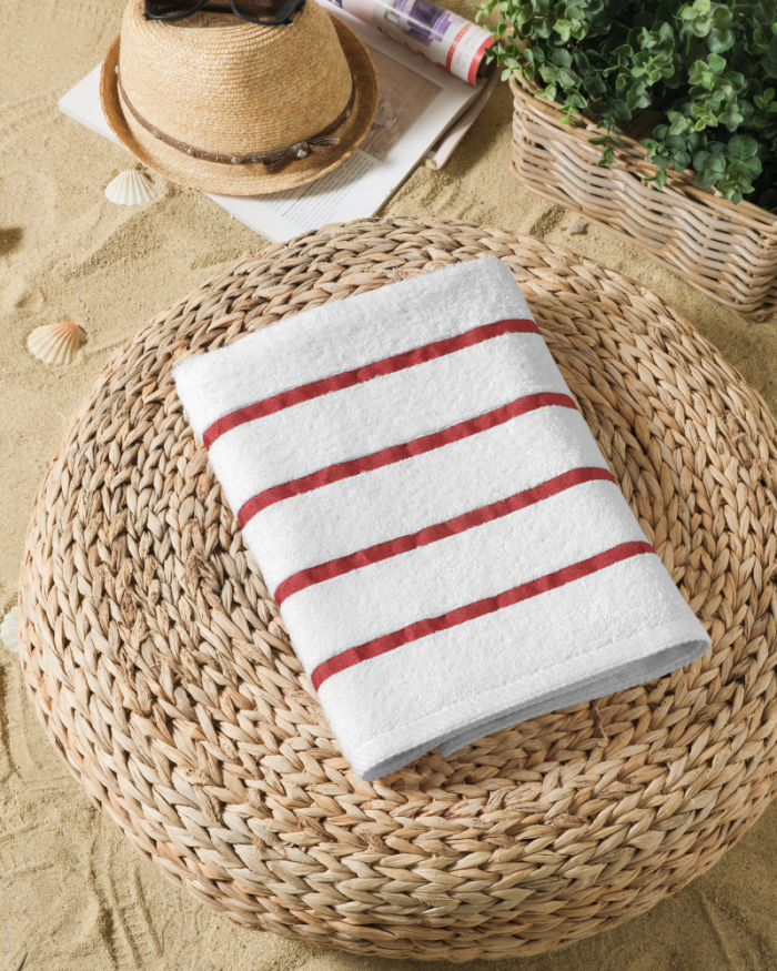 White towel with red stripes on a woven mat, beside a straw hat and greenery on a sandy surface.