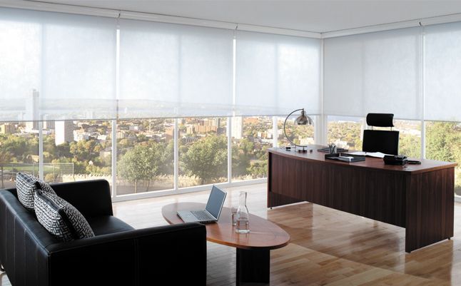 Modern office with large windows, a black sofa, coffee table, and wooden desk overlooking a city.
