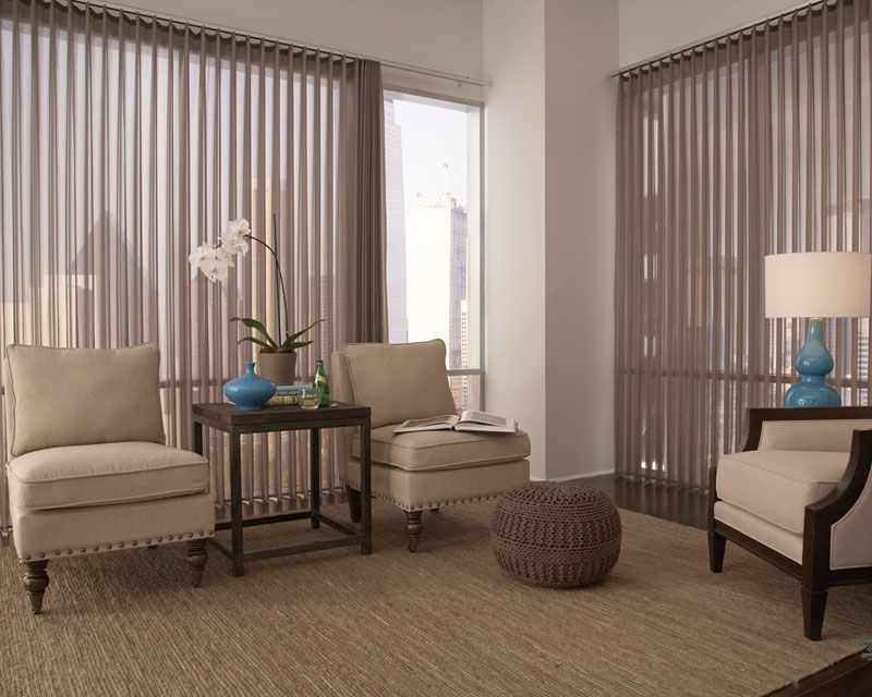 Modern living room with beige chairs, dark side tables, floor-to-ceiling blinds, and a blue vase centerpiece.