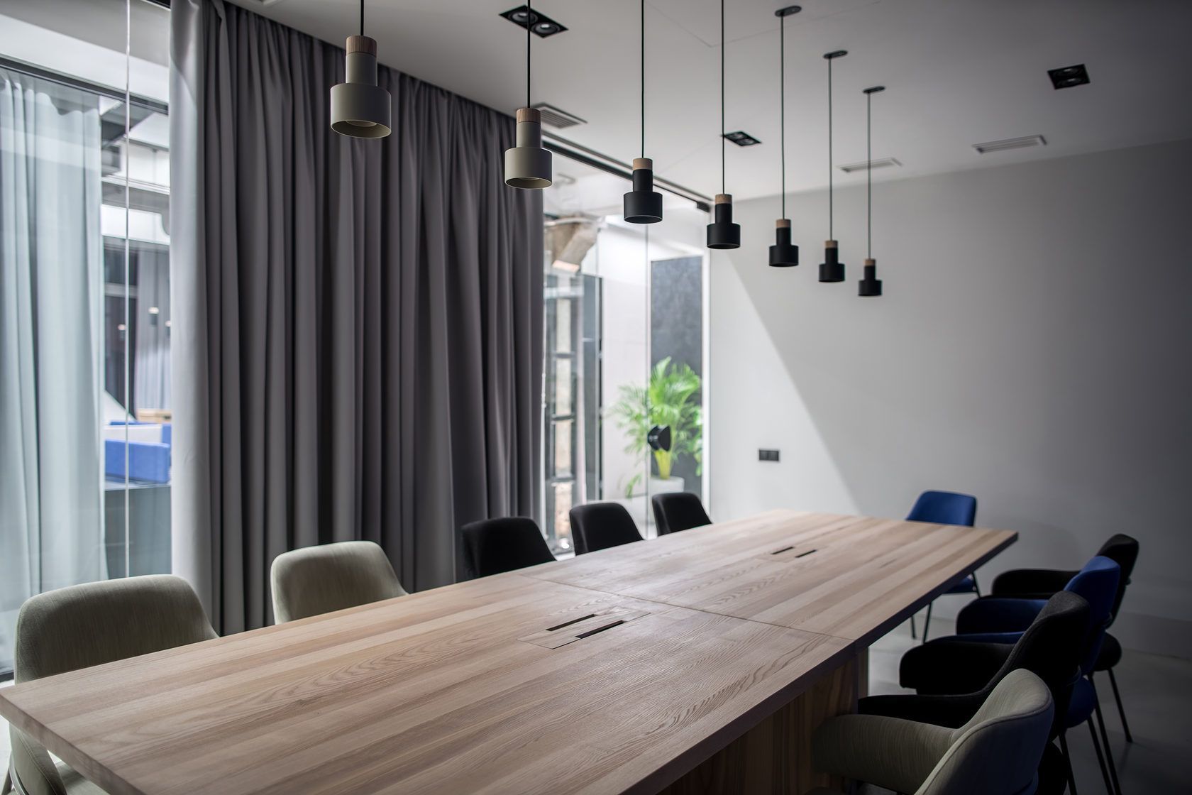 Modern meeting room with long wooden table, black chairs, gray curtains, and hanging pendant lights.