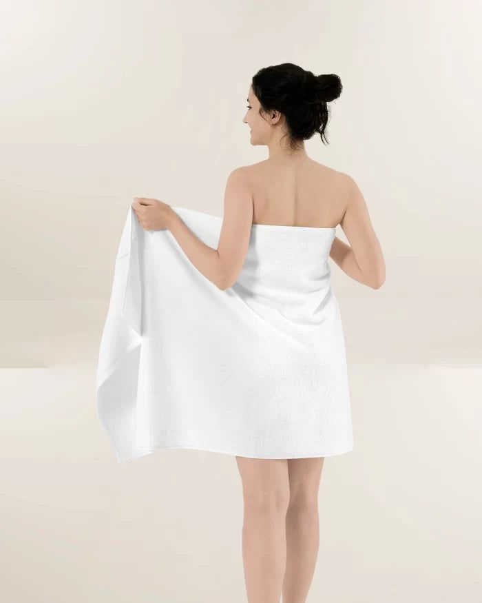 Person wrapped in a white towel, standing with back turned against a plain light background