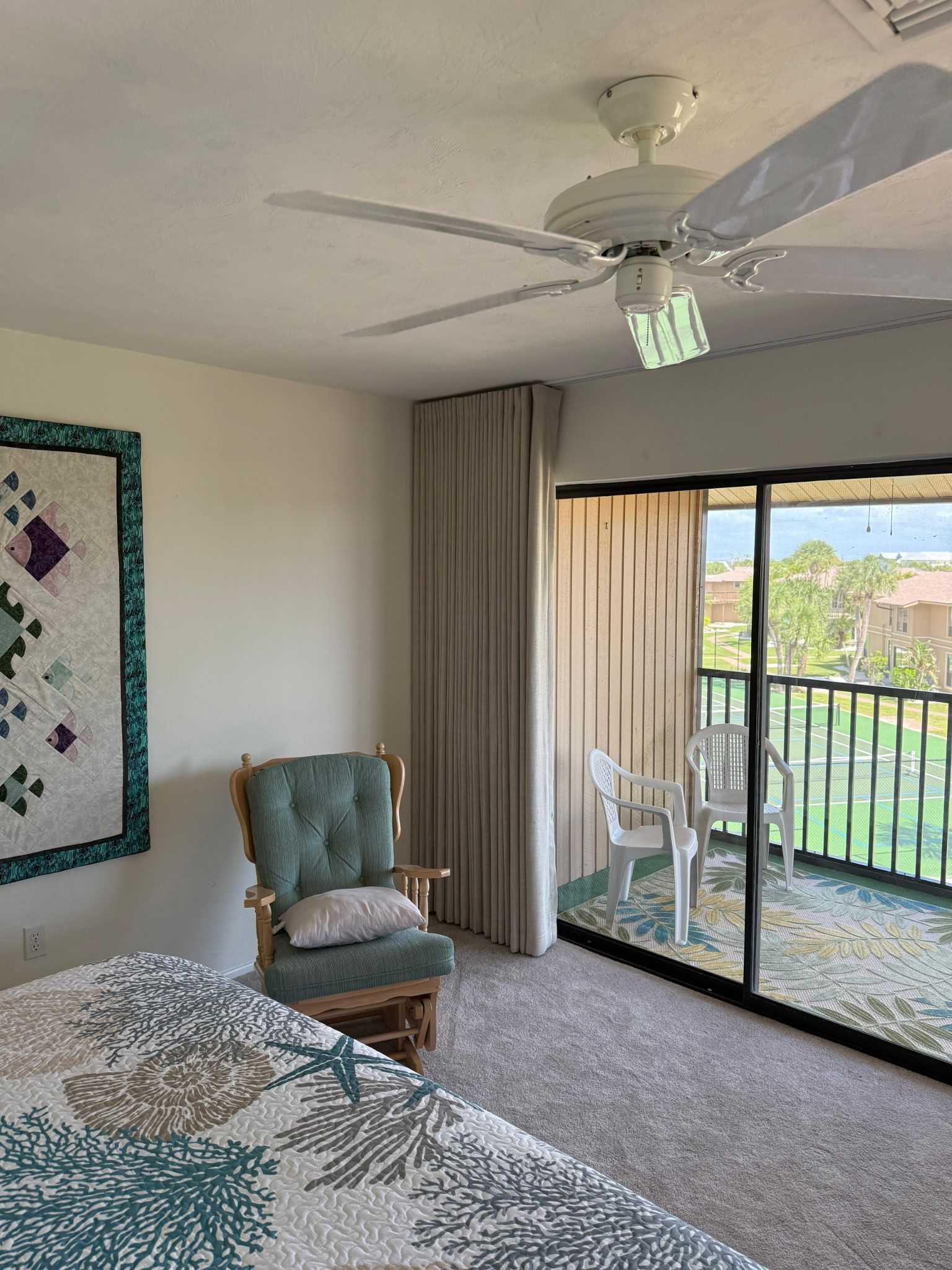 Bright bedroom with ceiling fan, quilted bed, armchair, and open sliding door to a balcony with mountain view.
