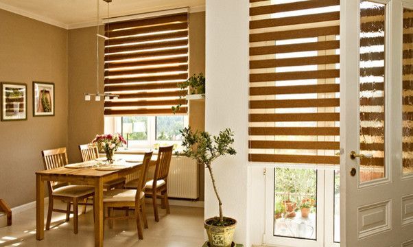 Bright dining room with wooden table, chairs, potted plants, and brown striped blinds by tall windows