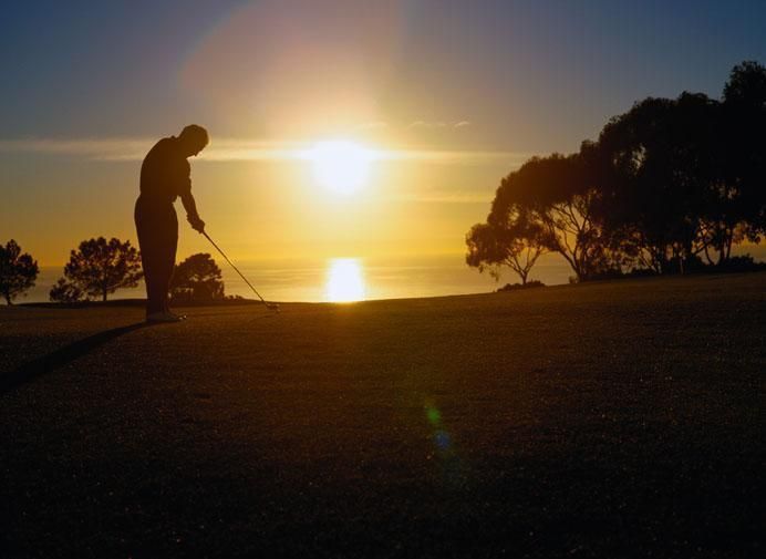 Golfer silhouetted against a sunset over the ocean, preparing to swing on the green.