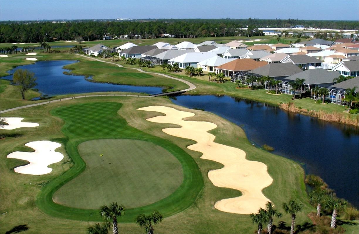 Aerial view of a golf course with green grass, sand traps, a lake, and houses in the background.