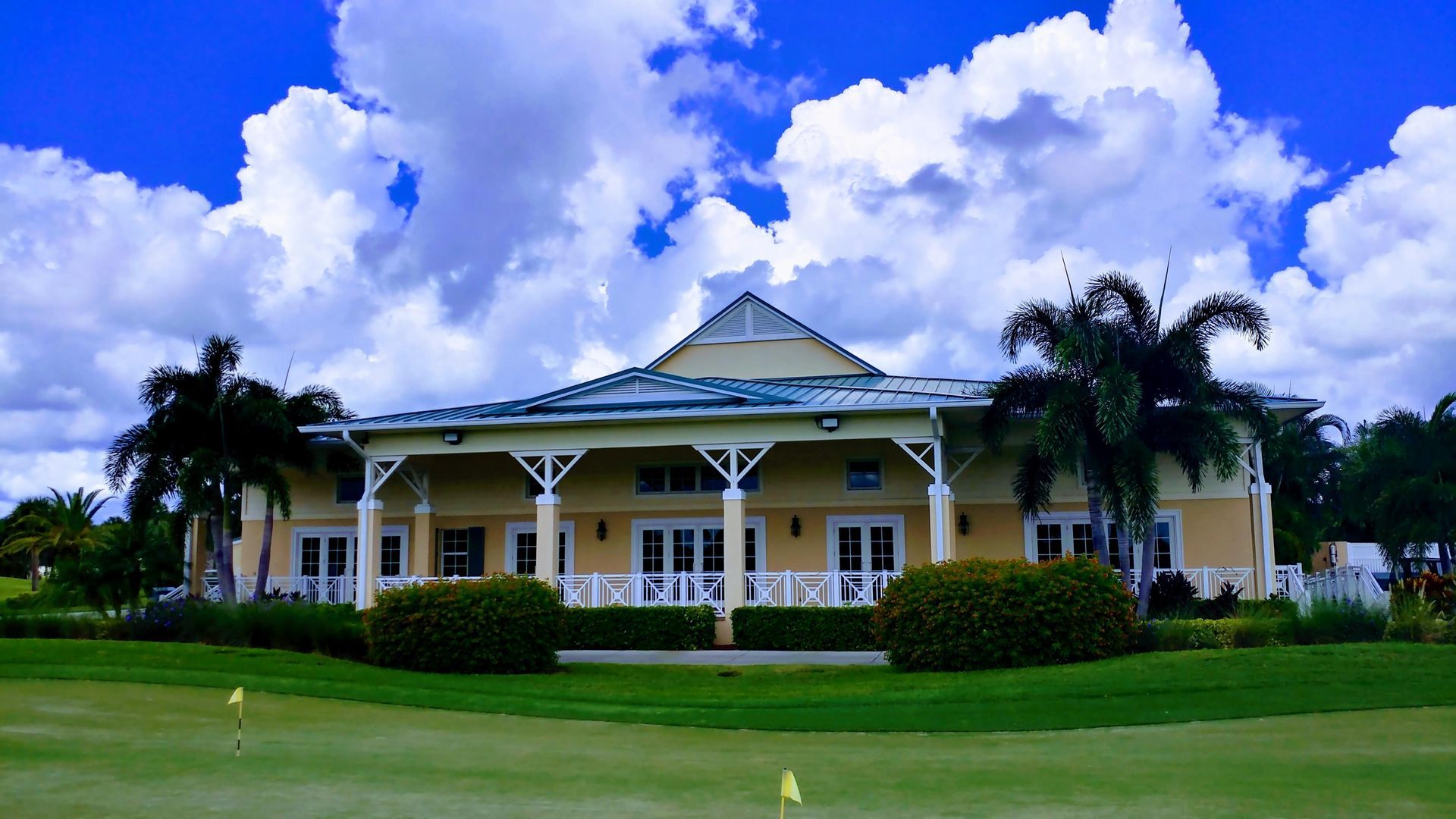 Yellow building with porch, palm trees, and green grass against a blue sky with clouds.