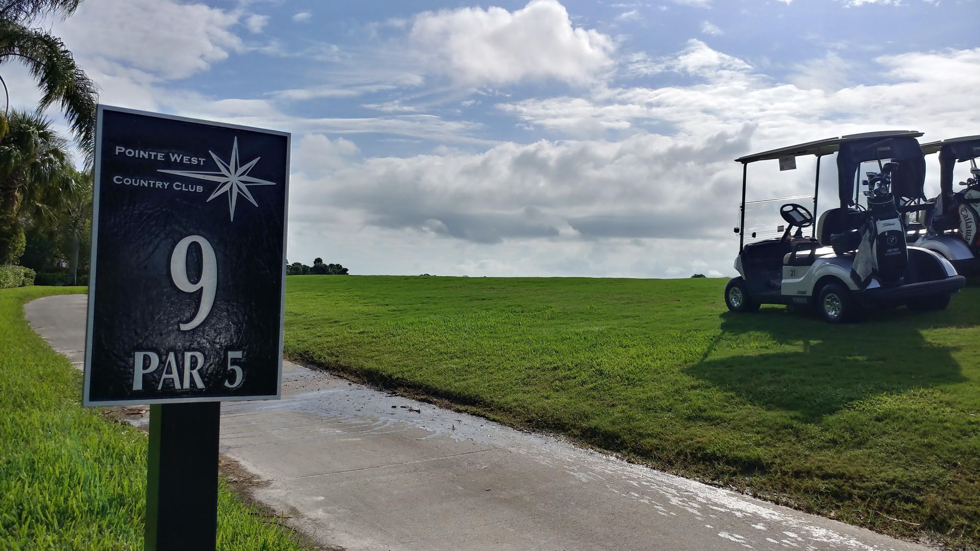 Sign for hole 9, par 5 at a golf course, with golf carts parked on a grassy field under a cloudy sky.