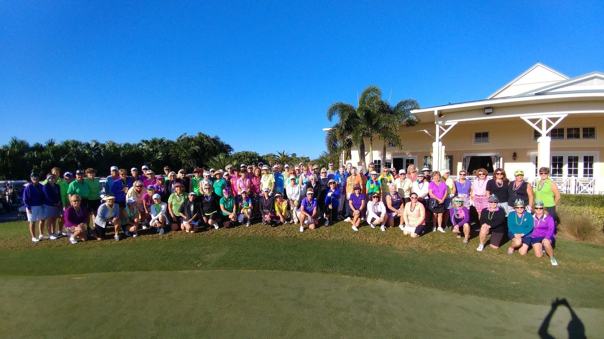 Large group of people in colorful clothes posing on a green golf course, with a building and palm trees in the background.