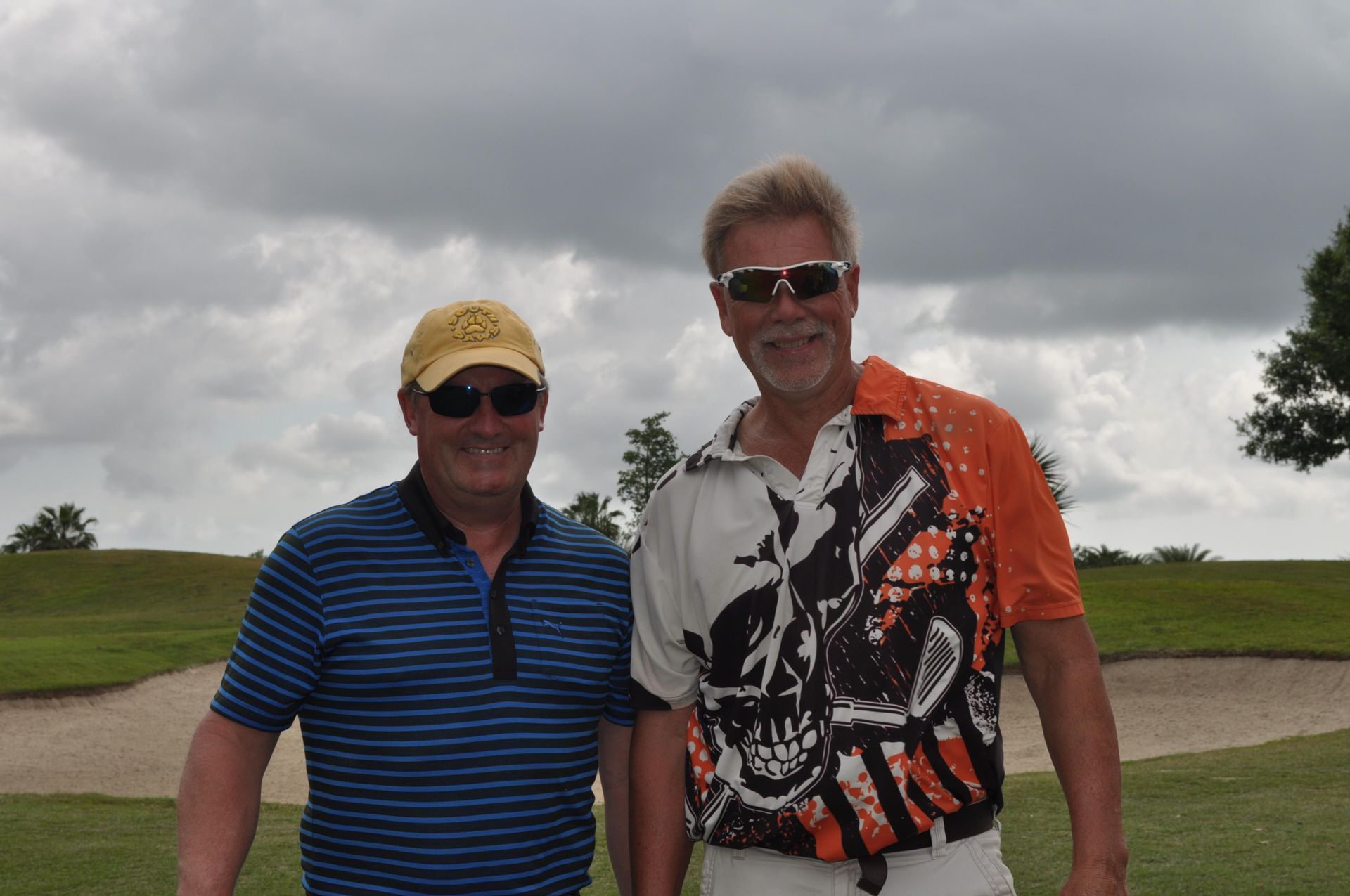 Two men on a golf course, one in a yellow hat and striped shirt, the other in sunglasses and an orange shirt.