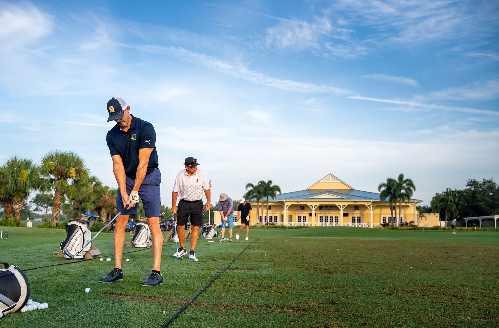 Golfers practicing on a driving range, with a clubhouse in the background under a blue sky.