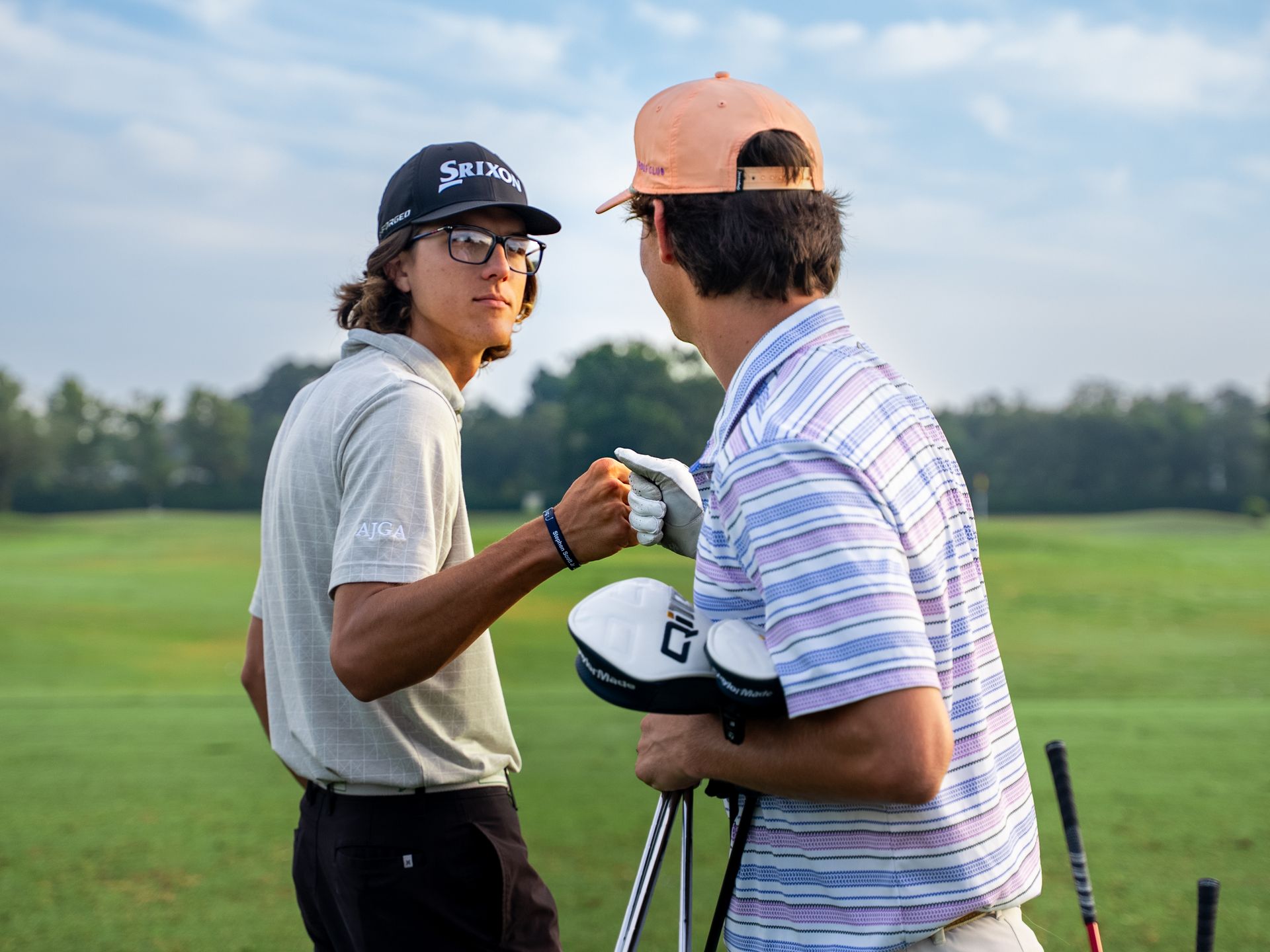 Two golfers fist bump on a green golf course, one wearing a hat and glasses, the other holding golf clubs.