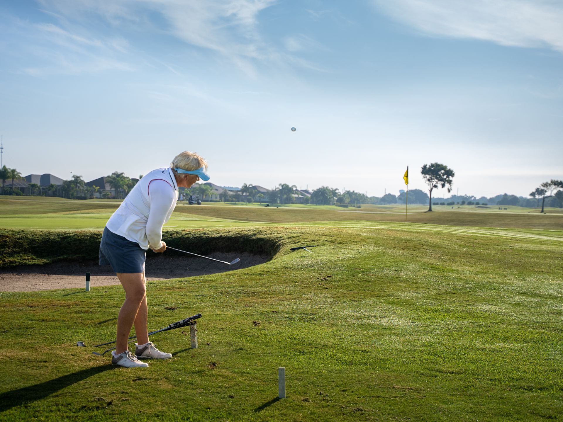 Woman swings golf club on sunny green course. Ball in flight, flag in distance.
