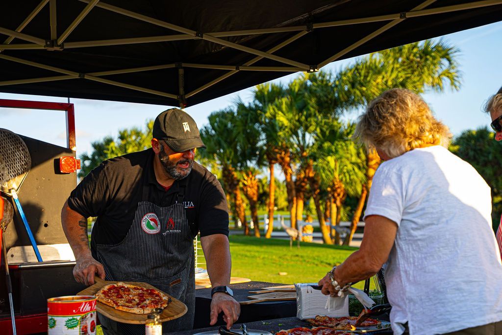 Man serving pizza to a customer at an outdoor event, palm trees in the background.