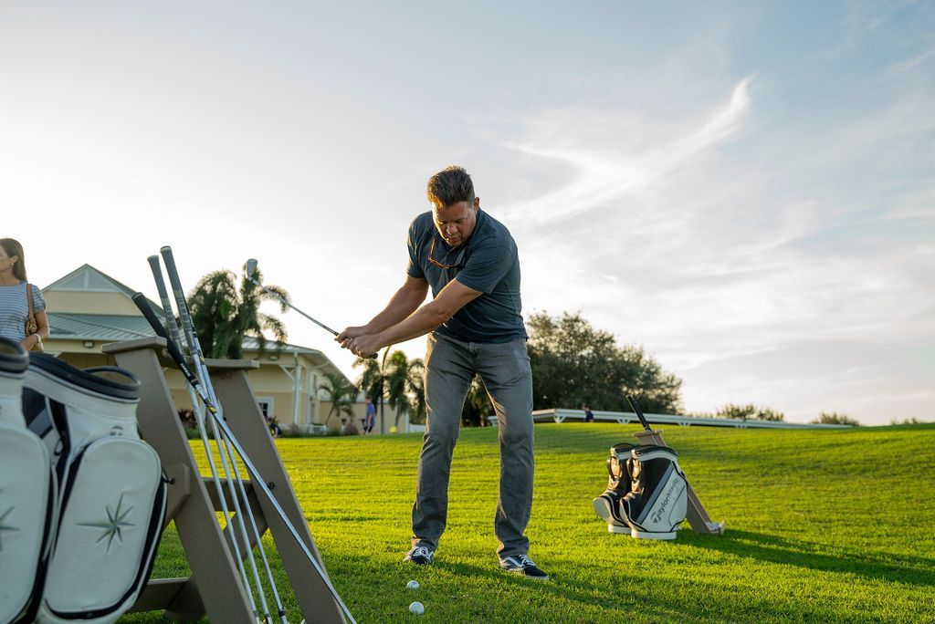 Man swinging a golf club on a green course with golf bags and balls. Bright sunlight.