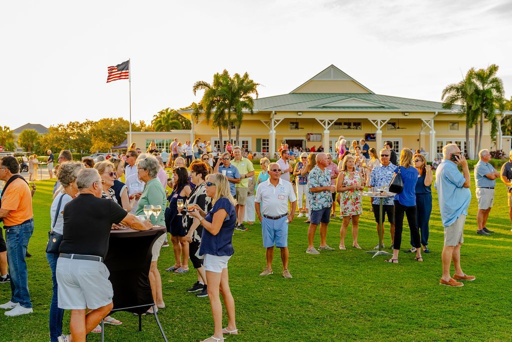 Large outdoor gathering with people socializing on a lawn in front of a building; US flag in background.