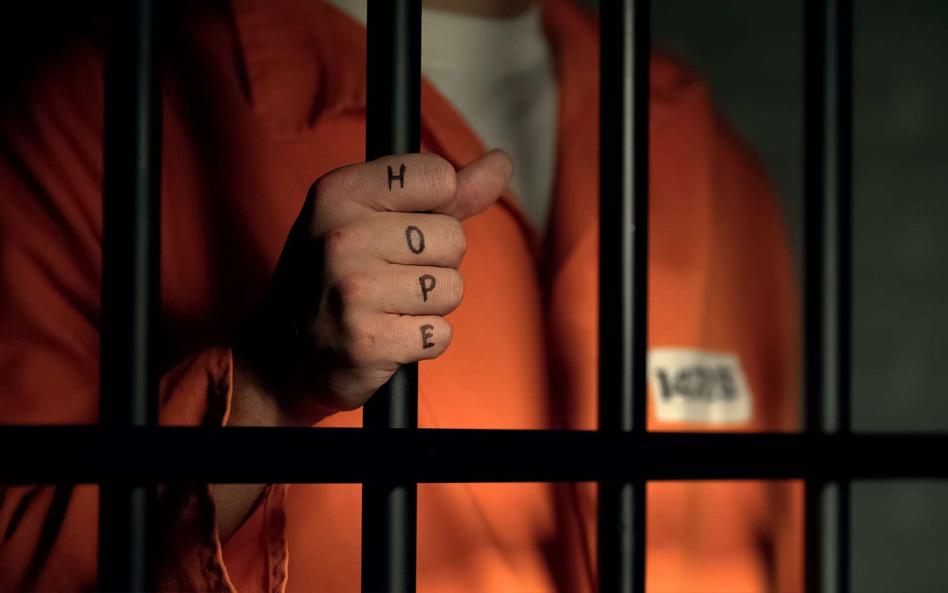 A man in an orange jumpsuit is behind bars in a prison cell.