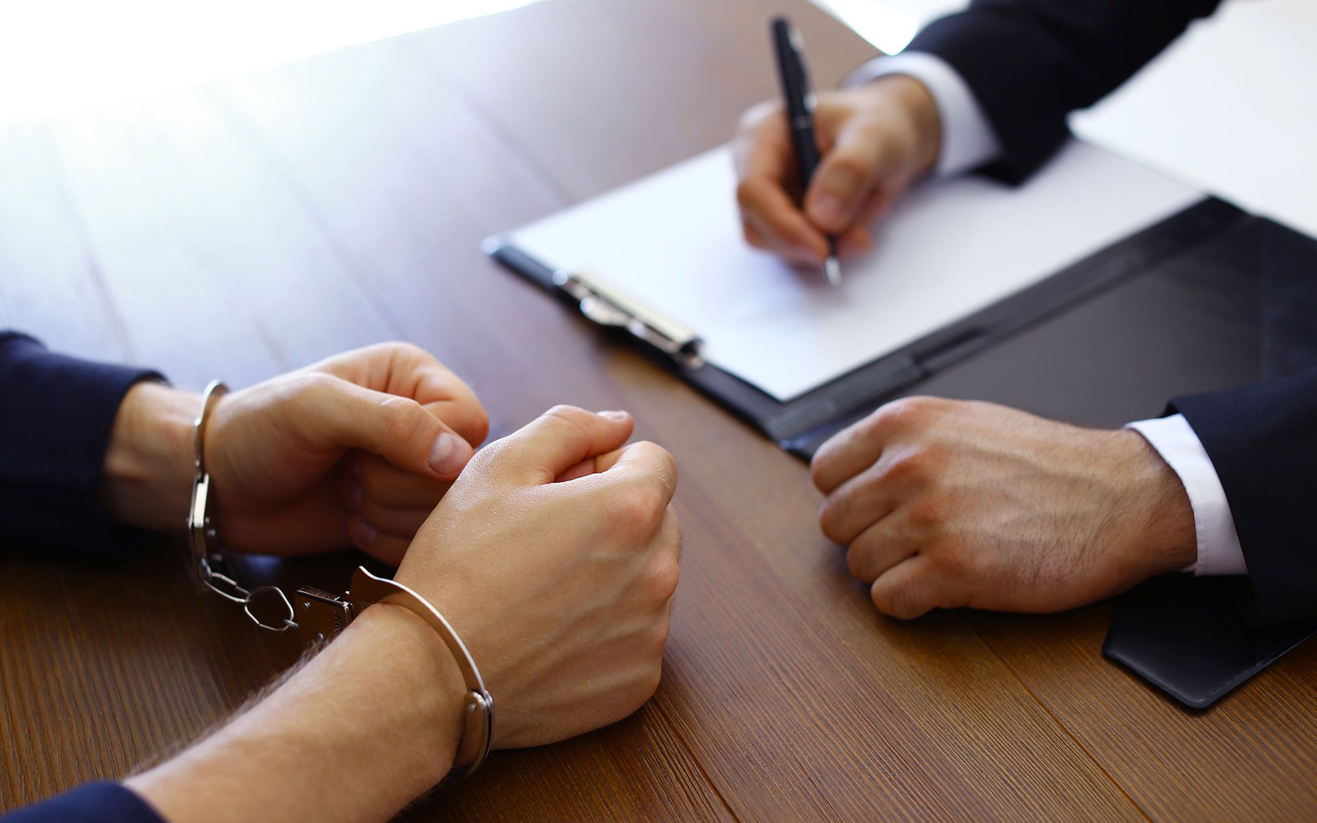 A man in handcuffs is sitting at a table with a man writing on a clipboard.