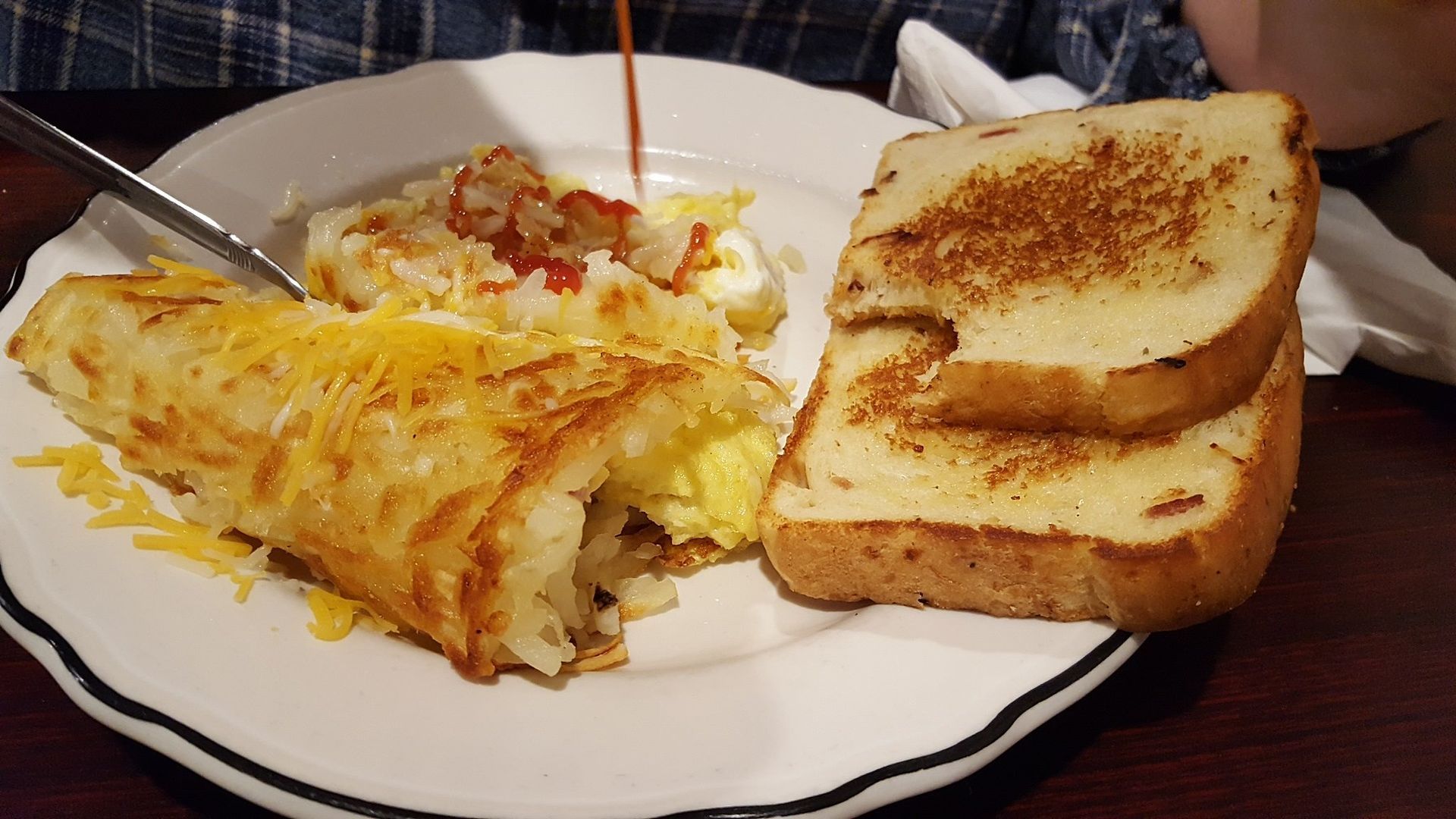 A plate of food with eggs , potatoes , toast and syrup being poured on it.