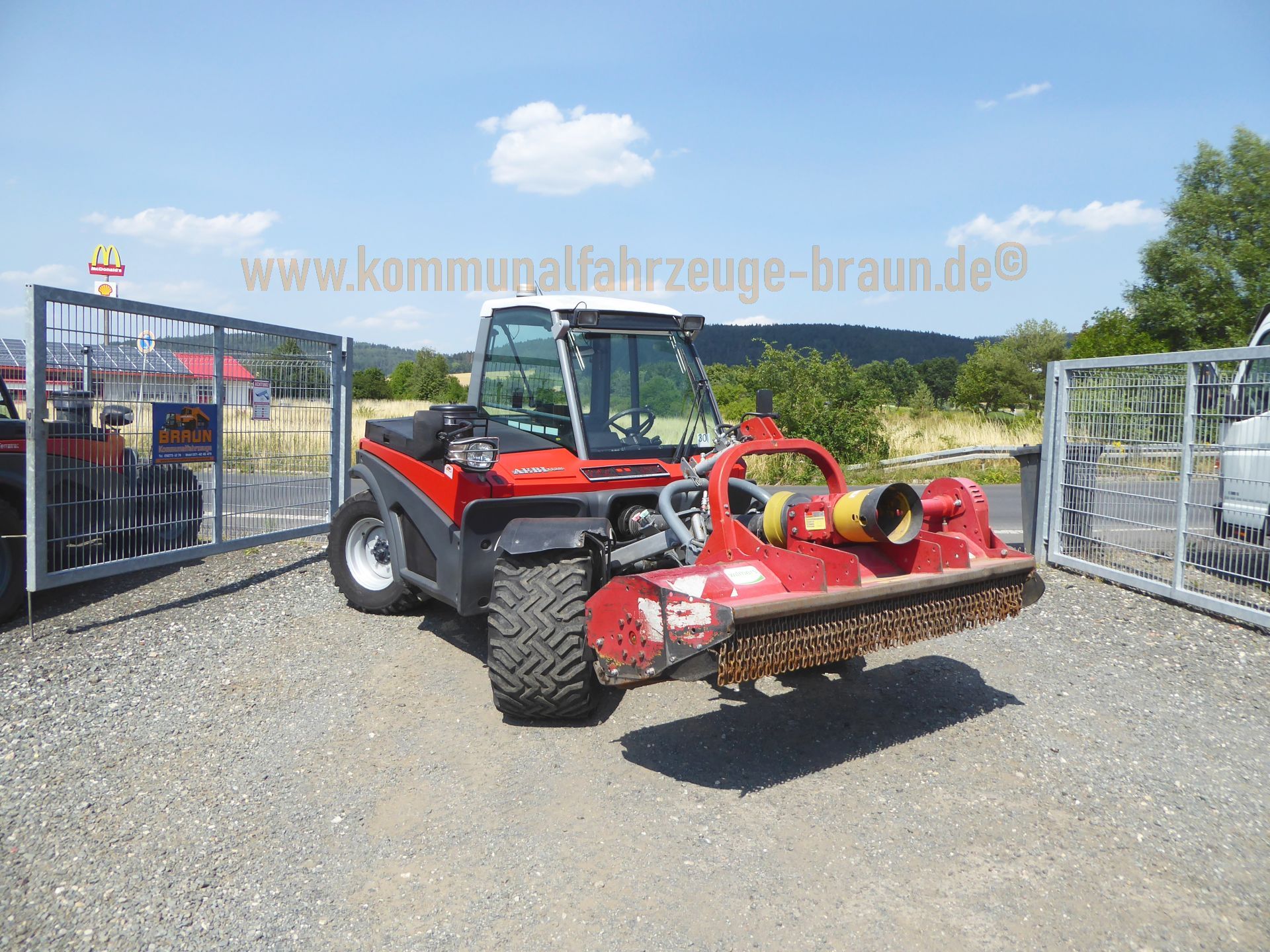 A red tractor is parked in a gravel lot next to a fence.