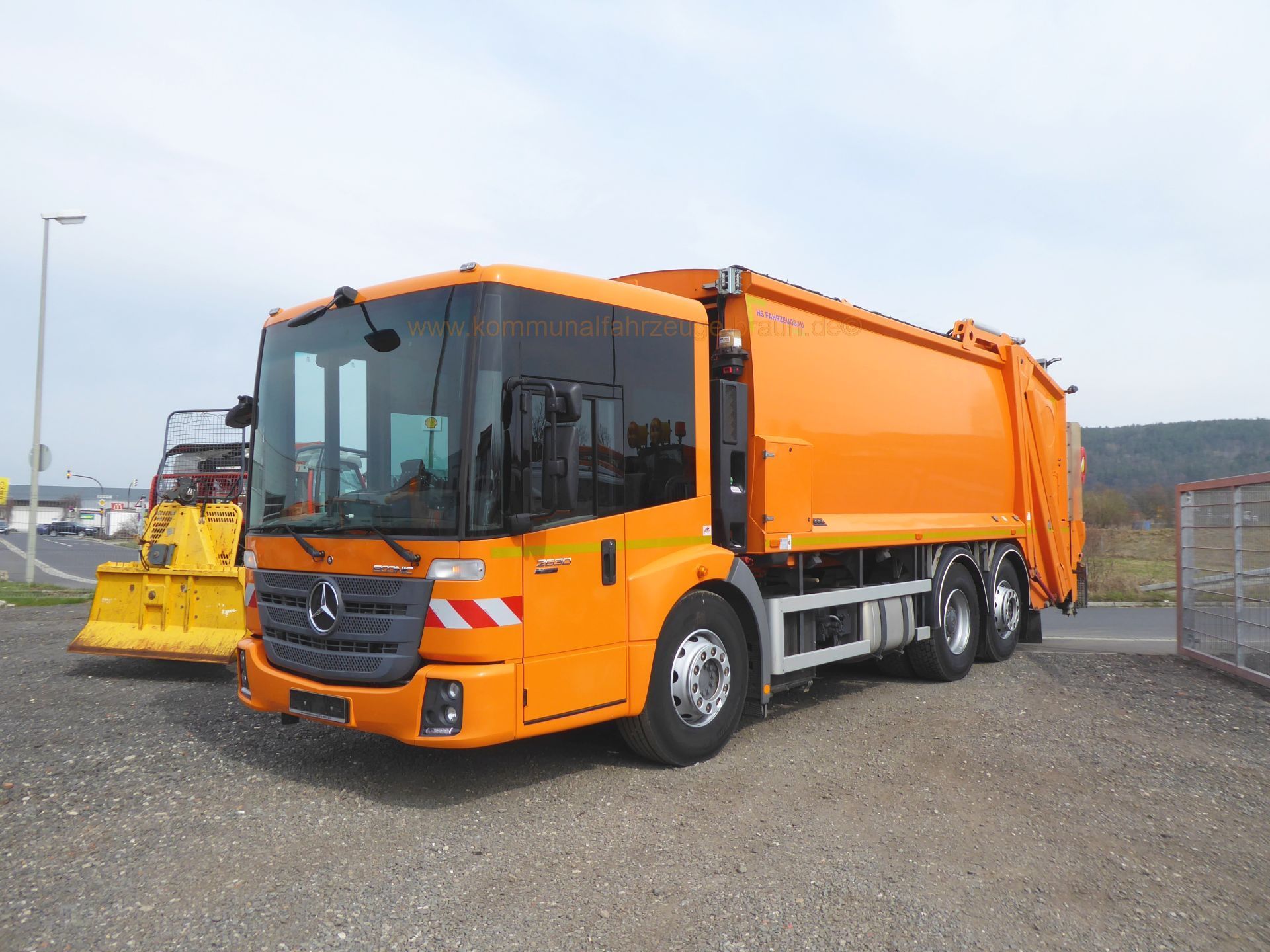 An orange garbage truck is parked in a gravel lot.