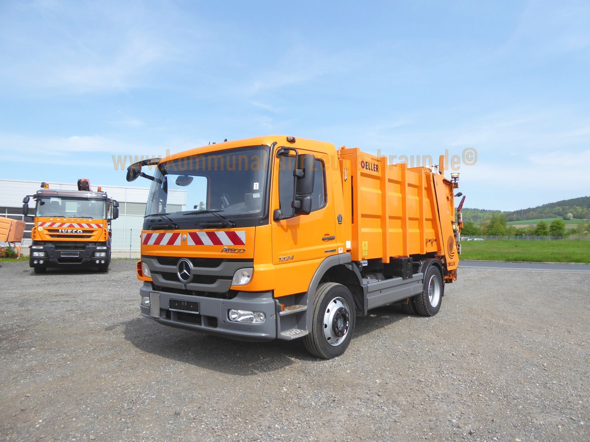 Two orange garbage trucks are parked in a gravel lot.