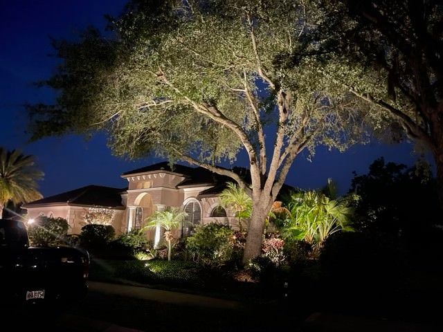 Night view of a house with landscape lighting, illuminating the facade and large trees, blue sky in the background.
